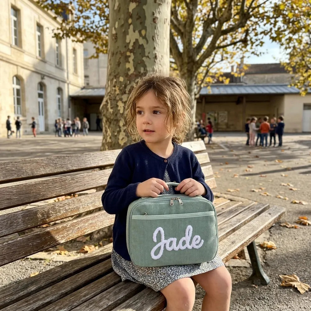 Petite fille assise sur un banc de cour d'école avec son sac à goûter vert sauge brodé Jade